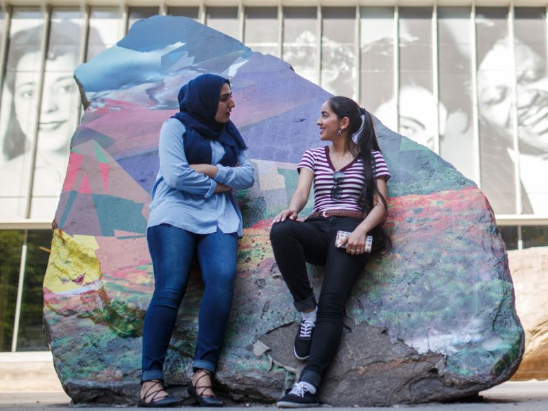 Two students chat in front of a painted rock on TMU's campus