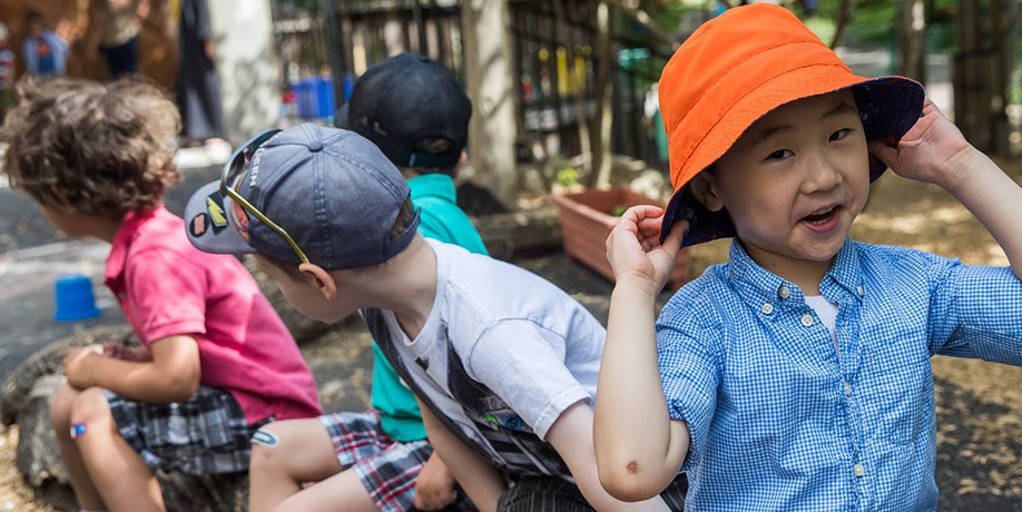 Young children in an outdoor playground in summer; a boy in a checked shirt smiles and holds his orange hat