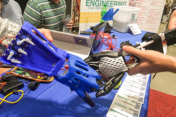 Two students hold prototypes of prosthetic arms with their hands interlocked.