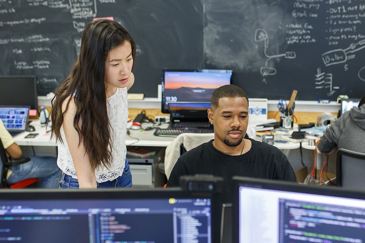 Inside a computer lab with chalkboards on the wall, two students work together on a desktop computer. 