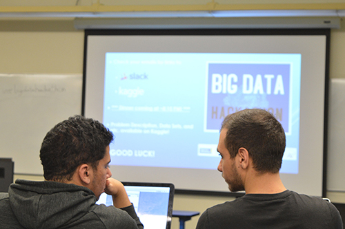 Two students sit in a classroom and face a large screen displaying information about a Big Data Hackathon.