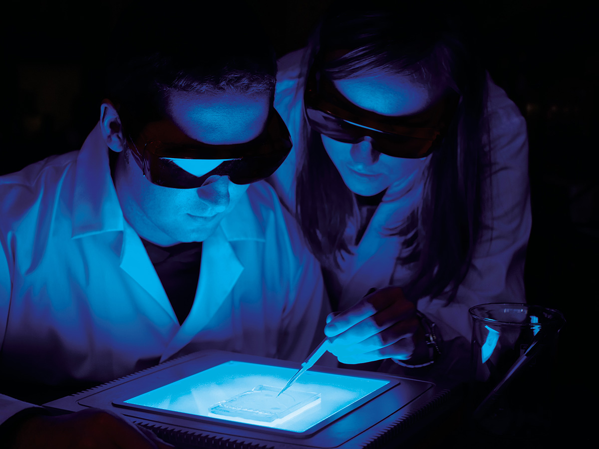 Two students in safety glasses and white lab coats examine a brightly lit surface.