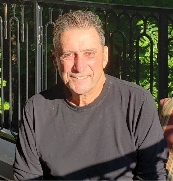 A man in a black shirt smiles while sitting outdoors in bright sunlight. In the background, there's a black metal railing and lush green foliage.