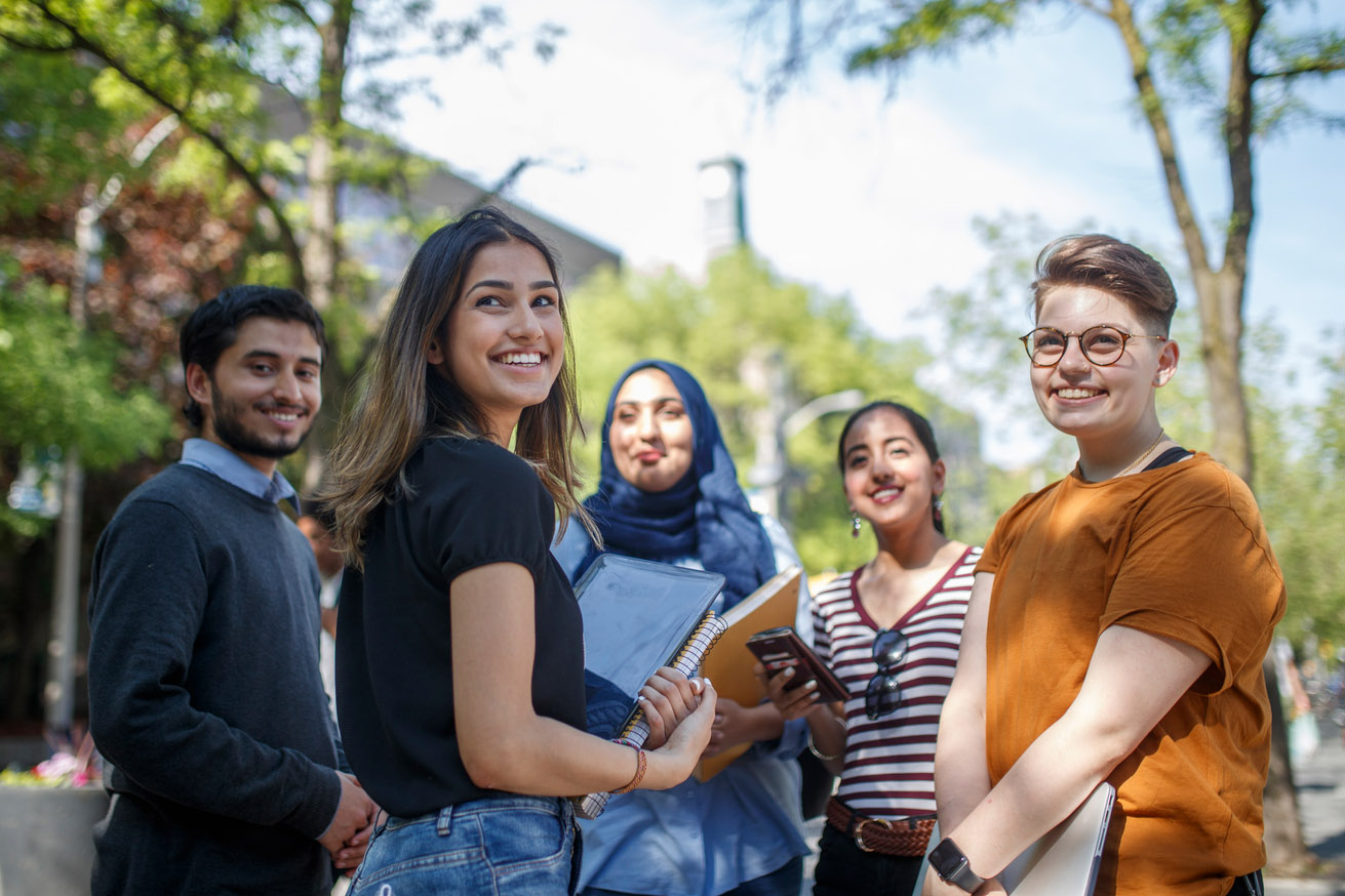 A group of five smiling students stand together in a circle outside on the Toronto Metropolitan University campus.