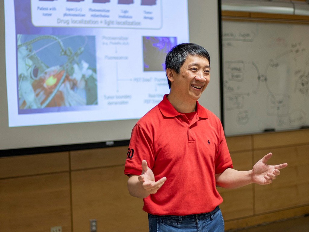 Prof. Victor Yang stands in a lecture hall with a smile, animatedly presenting with a screen projection behind