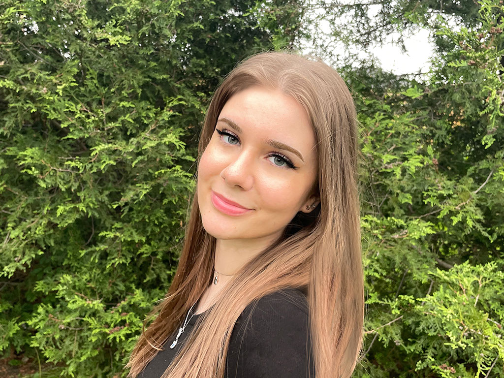 Jana Stojanovska looks at the camera, standing in front of a tall green hedge