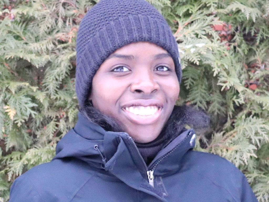 Jayda Brown, DAS graduate, smiles at the camera in front of a green hedge, wearing a blue hat and jacket