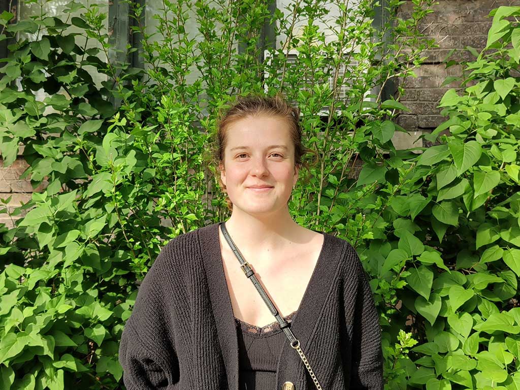 Leandra Budau smiles at the camera standing in front of green plants