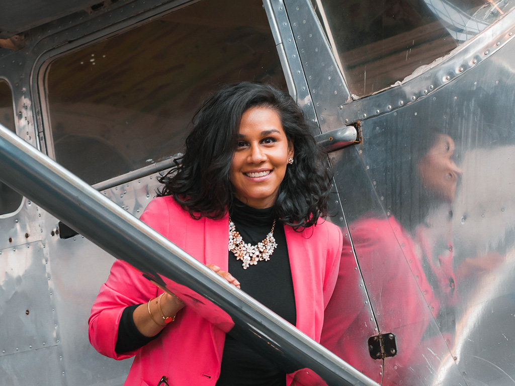 Sabrina Gomez Vila stands smiling in front of a silver airplane wearing a pink sweater and black t-shirt