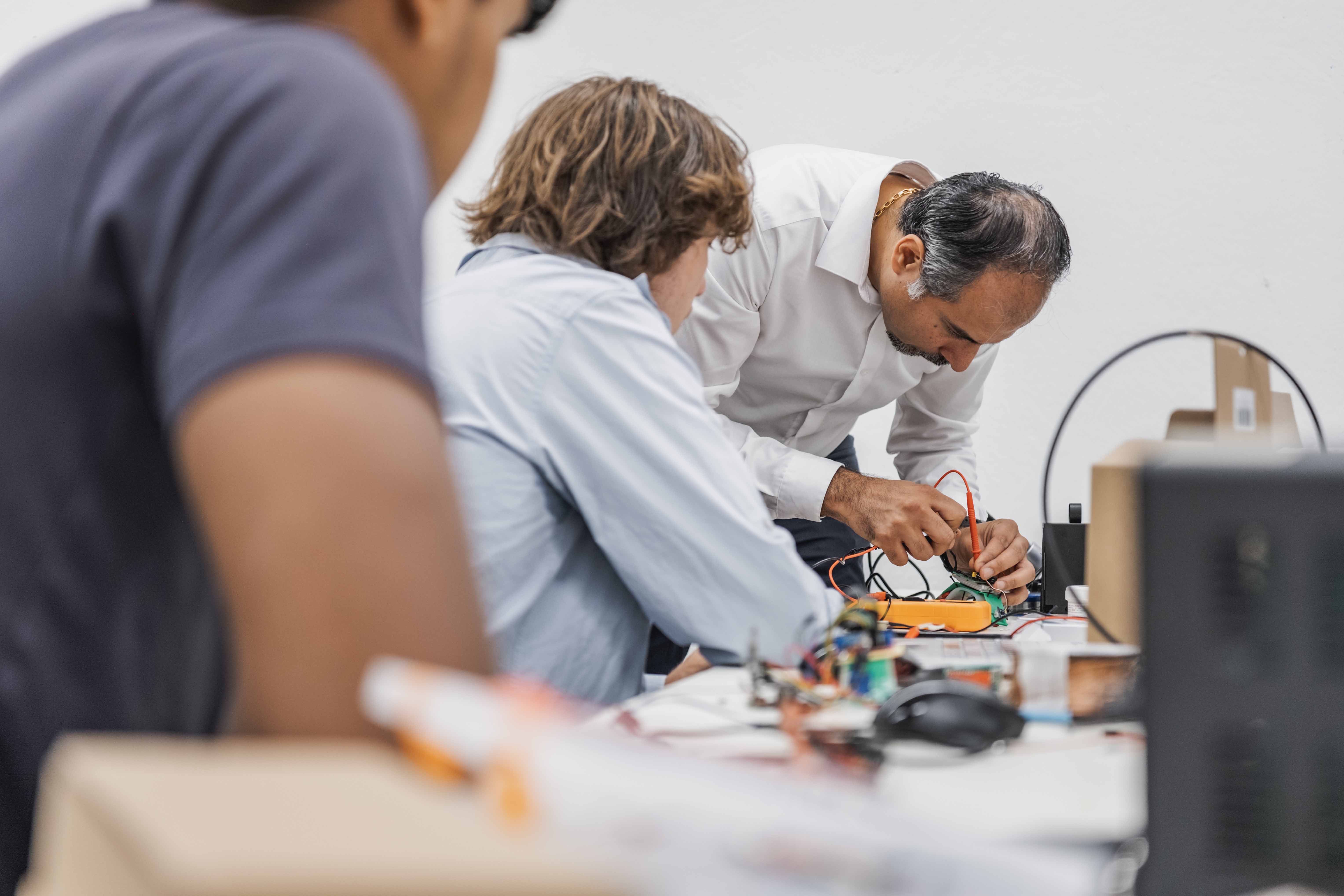 Professor Chhabra is configuring wiring for a robot next to two students in a lab with white walls. 
