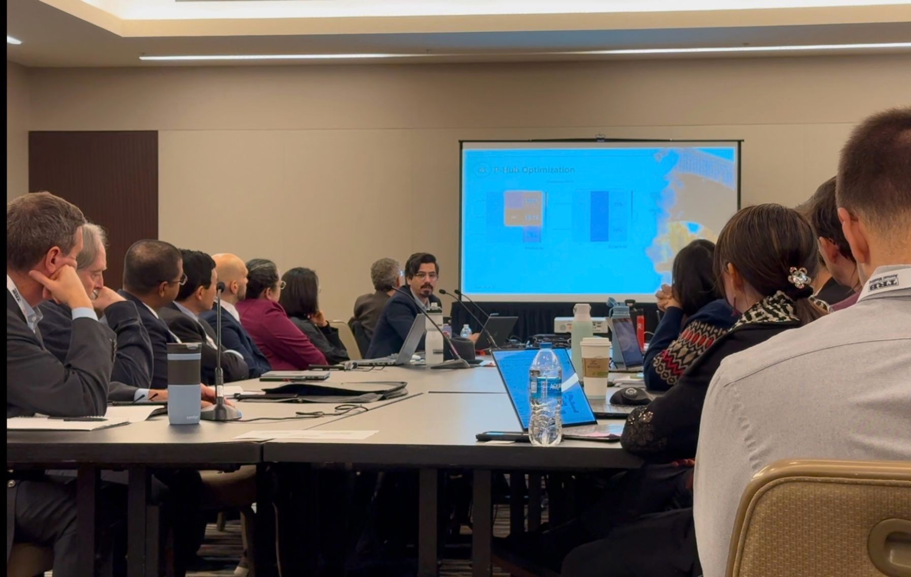 Alsaleh speaking with a group of people in a boardroom, with a presentation displayed  on a TV screen. 