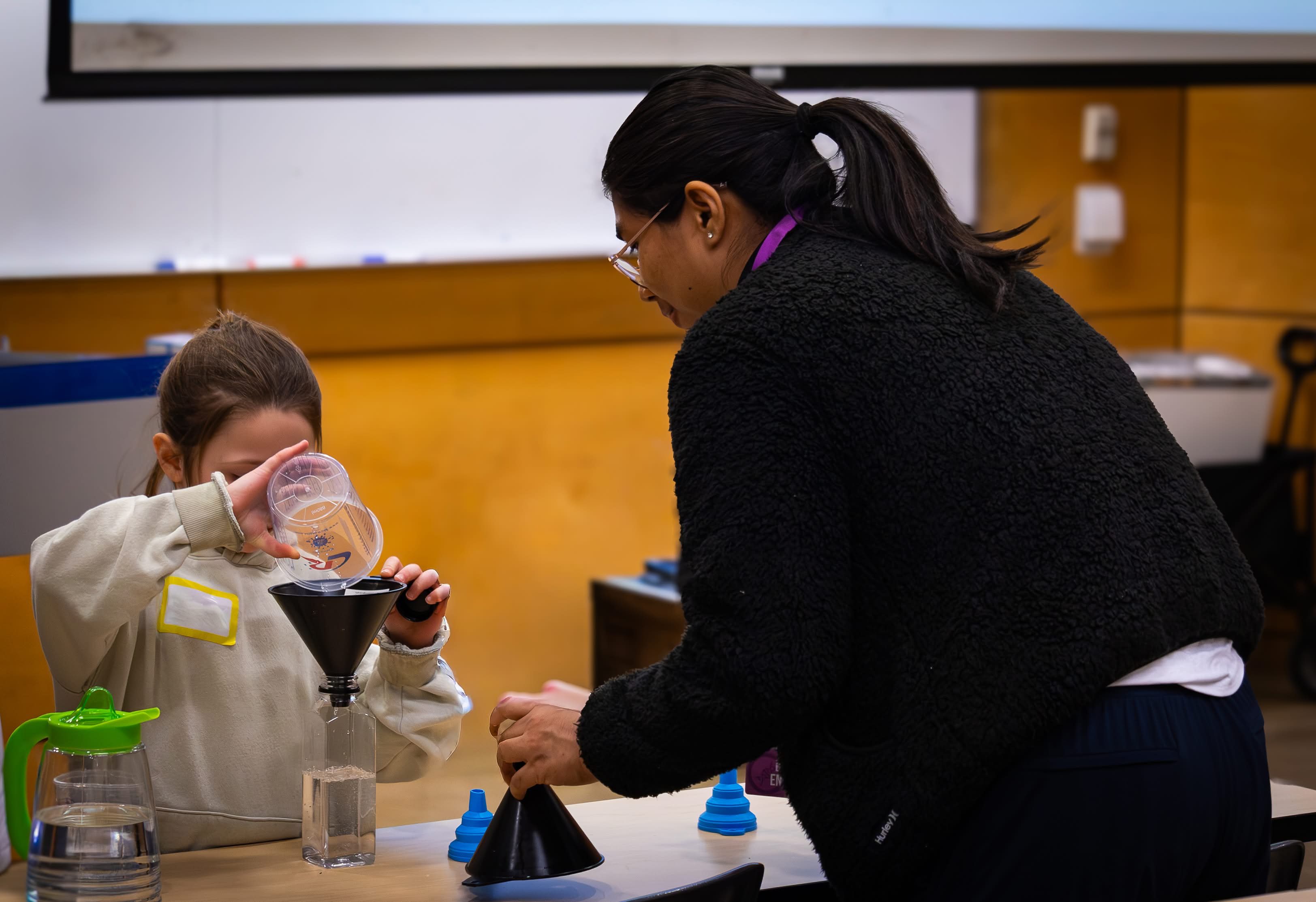 A student ambassador helps a Girl Guide pour liquid into a bottle using a funnel. 