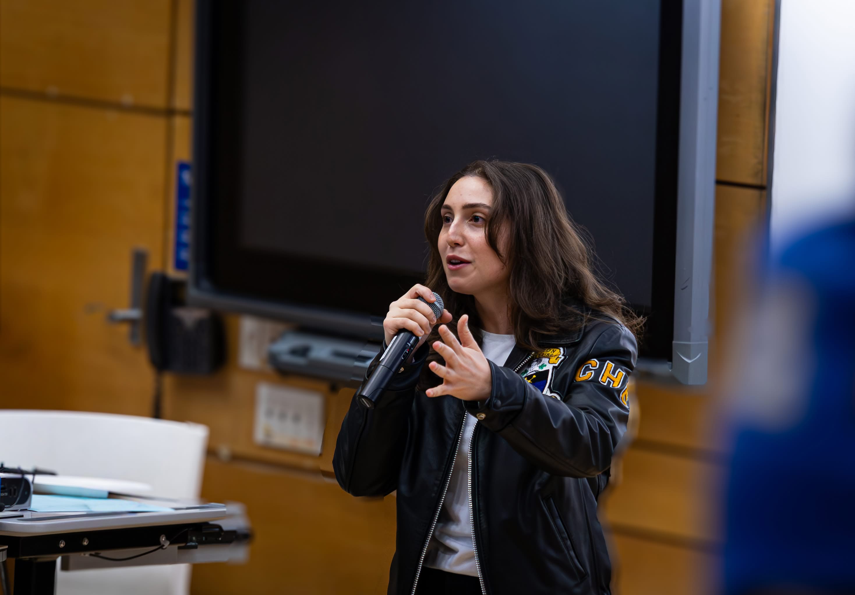 Young woman holding microphone and wearing a black embroidered jacket. 