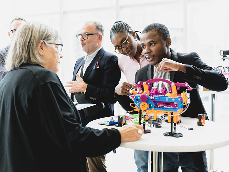 Naomi Adelson, Steven N. Liss and a few students stand near a table with a student’s colourful URO project on it