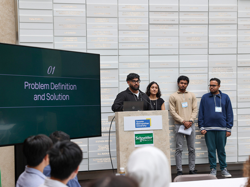 A student team presents their project beside a screen displaying "01 Problem Definition and Solution"