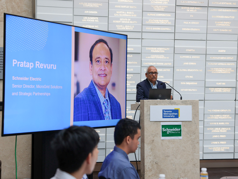 Pratap Revuru of Schneider Electric speaks at the podium beside a screen displaying his photo and title.