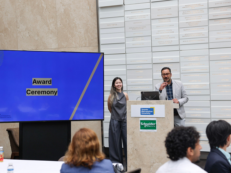 TMU staff members Mya Pham and Ashton Jila stand on stage, with Ashton speaking at the podium microphone during the award ceremony.