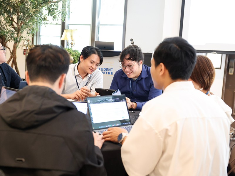 A group of participants sit together around a table, collaborating with laptops during an activity.