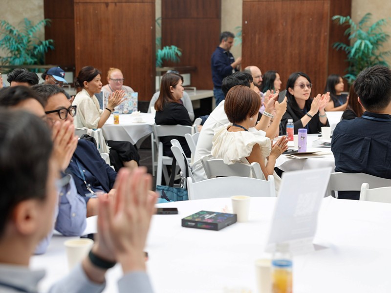 Audience members sit at tables and applaud during a presentation.
