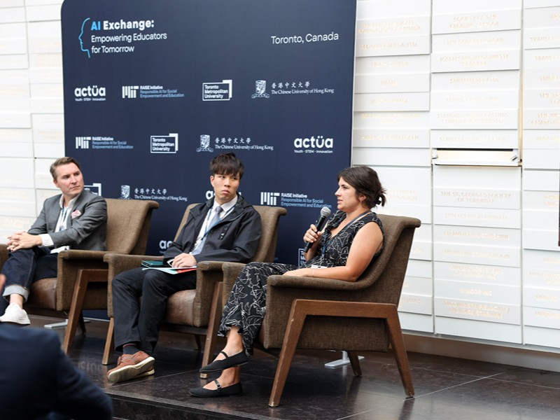 Three panelists sit on stage in front of the event backdrop while Dr. Emily Cyr holds a microphone and speaks.