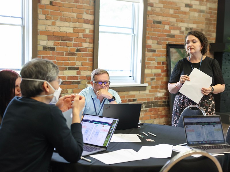 A speaker holds papers while addressing attendees gathered around a table for an activity.