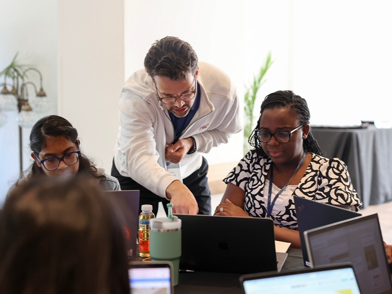  An activity leader leans over a table to guide two participants working on their laptops during a workshop session.