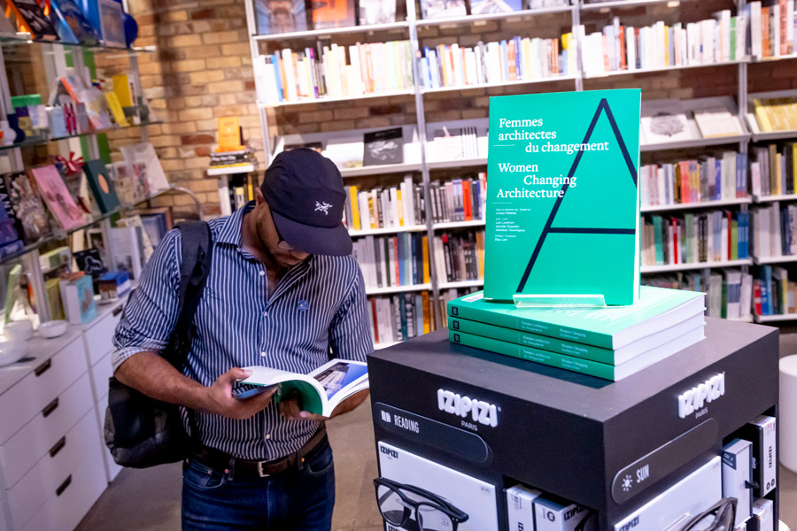 A man reading a copy of Women Changing Architecture next to a stack of green books.