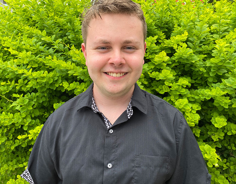 Joshua Jobe, the 2022 recipient of the Ontario Professional Engineers Foundation Education Gold Medal, stands in front of a green hedge smiling at the camera