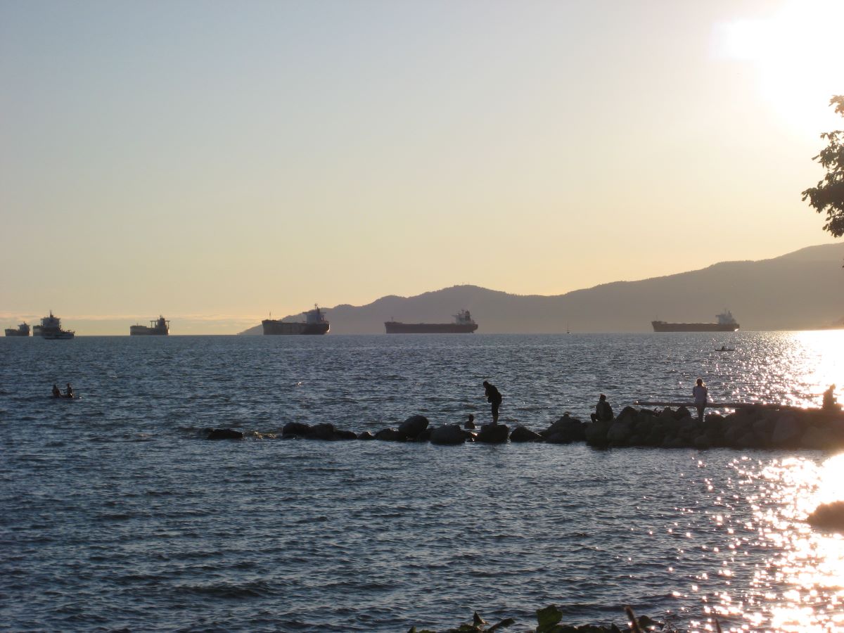 Burrard Inlet, Vancouver, British Columbia in Canada cargo boats pass by in the distance. People walk across rocks leading into the inlet. 