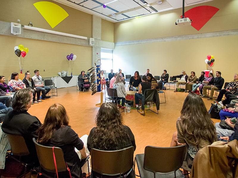 At the Gdoo-maawnjidimi Mompii Indigenous Student Services graduation, Indigenous and non-Indigenous community members gathered in a circle with drummers in the centre. The Eagle Staff stands nearby.