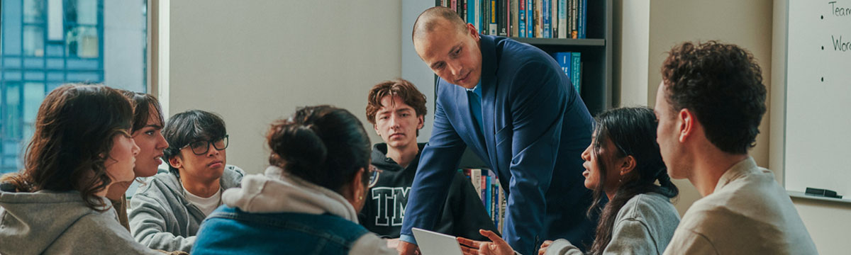 A professor among seven TMU students sitting at a table and listening to their peer who is chatting while looking at her open laptop.