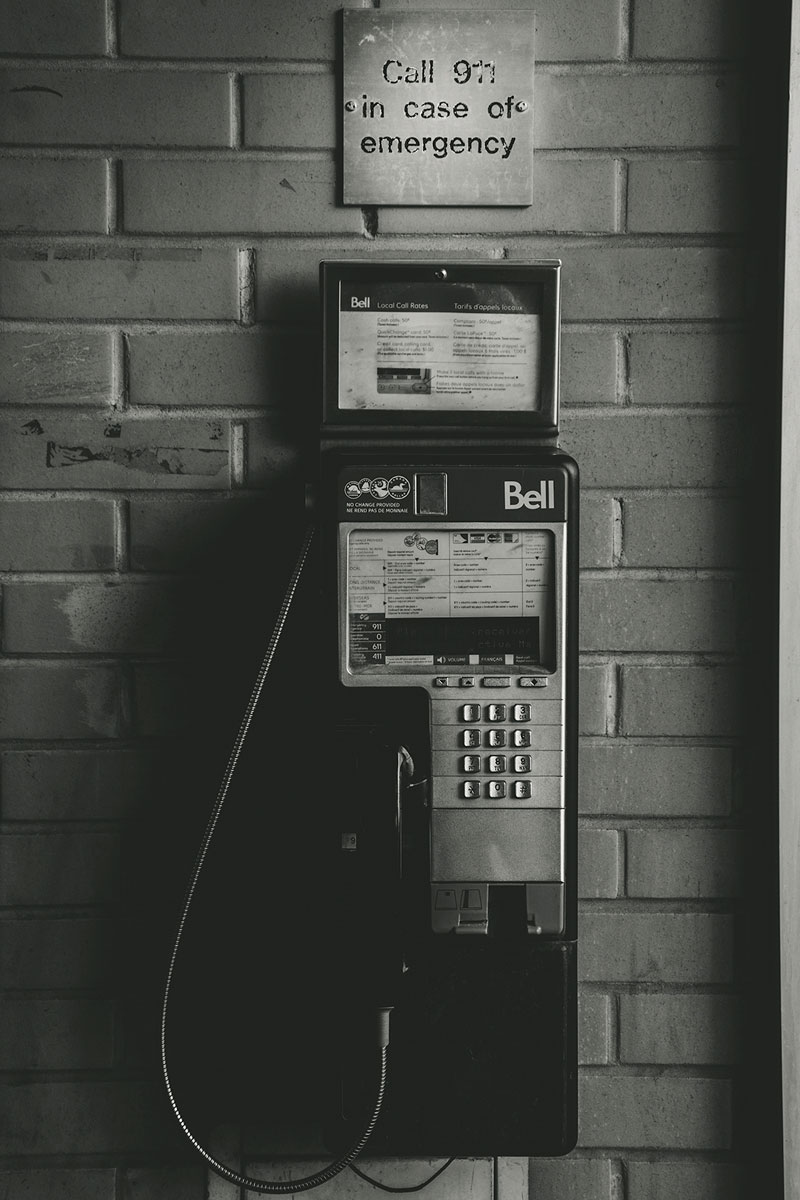 Payphone on a brick wall. A sign above it reads “Call 911 in case of emergency”