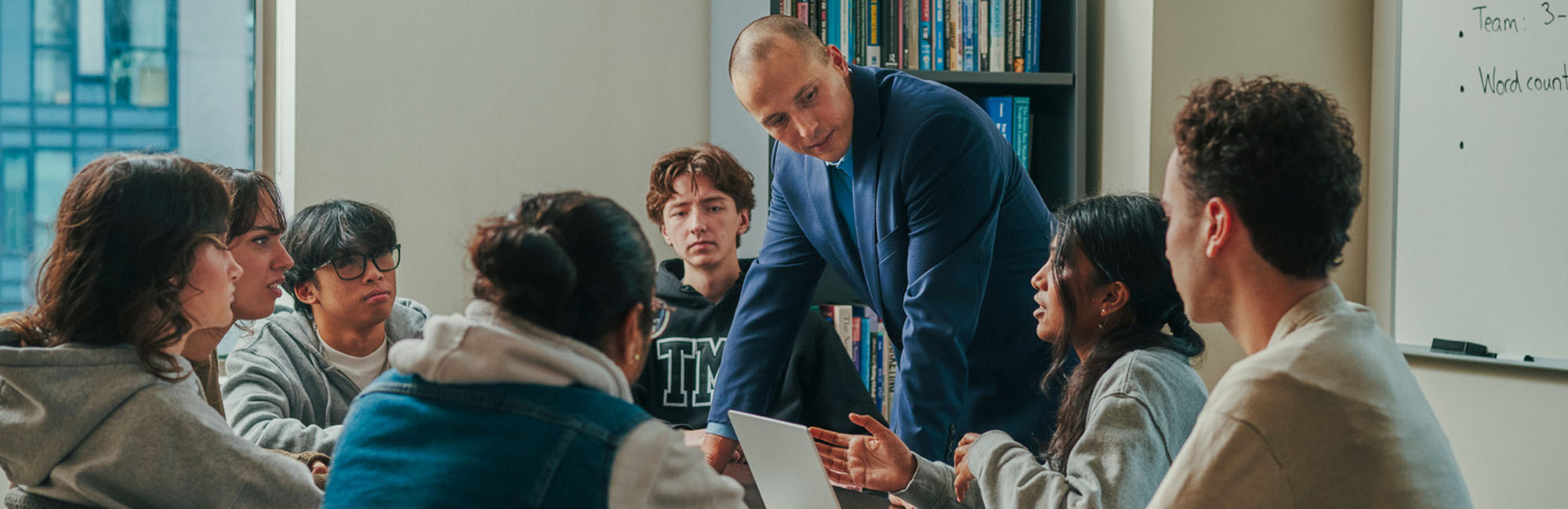 A professor among seven TMU students sitting at a table and listening to their peer who is chatting while looking at her open laptop.