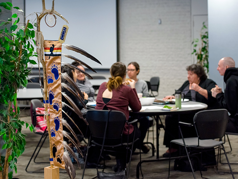 Elder Joanna Dallaire sitting with others around a table at TMU community consultations, the Eagle Staff stands nearby