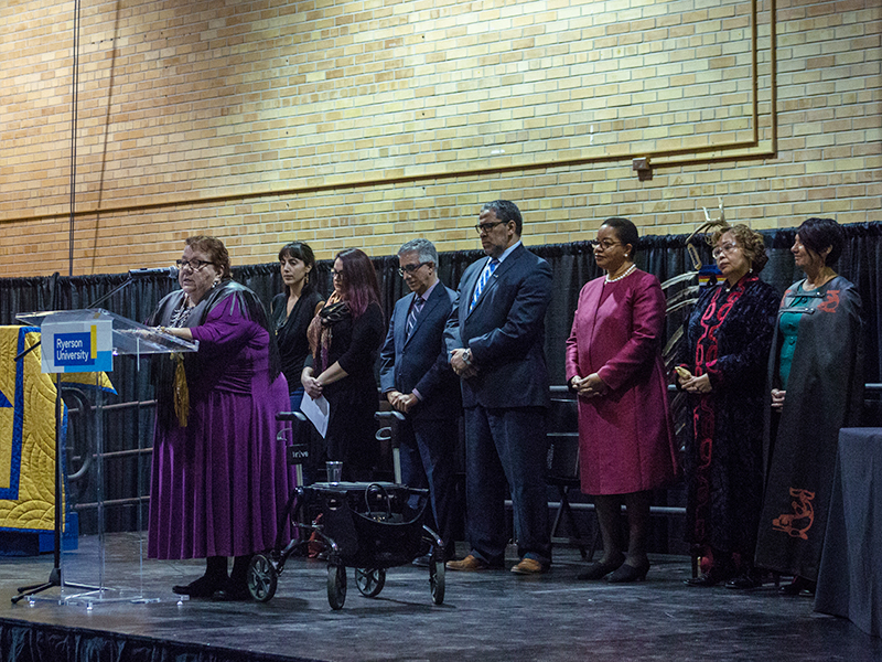 Elder Joanne Dallaire speaking at a podium at the Truth and Reconciliation at TMU community celebration; university leaders stand behind her in support