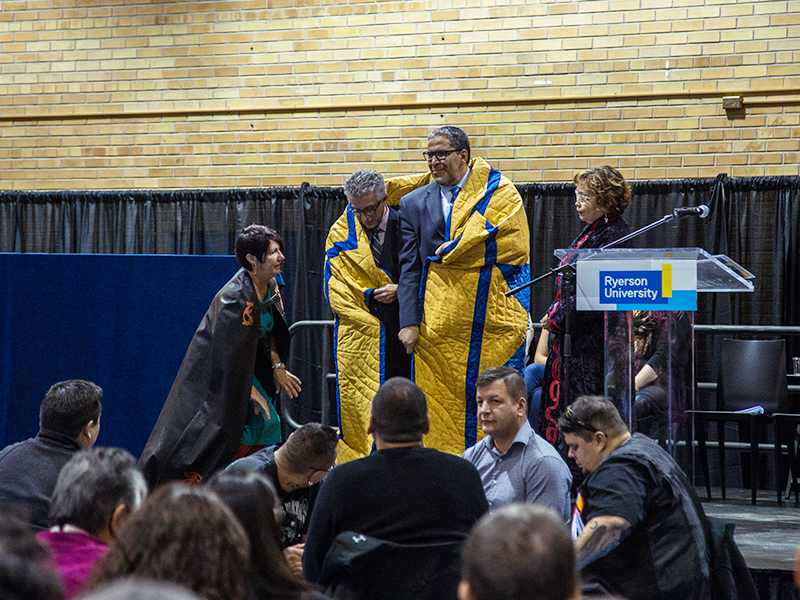 Michael Benarroch and Mohamed Lachemi being wrapped in the yellow and blue star blanket at the Truth and Reconciliation at Toronto Met community celebration