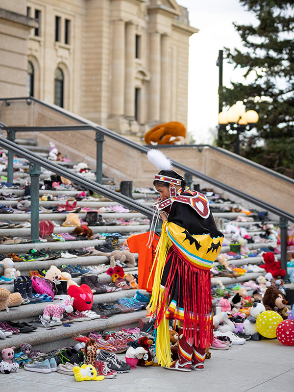 An Indigenous person dressed in indigenous regalia, looking at steps covered with children sized shoes 