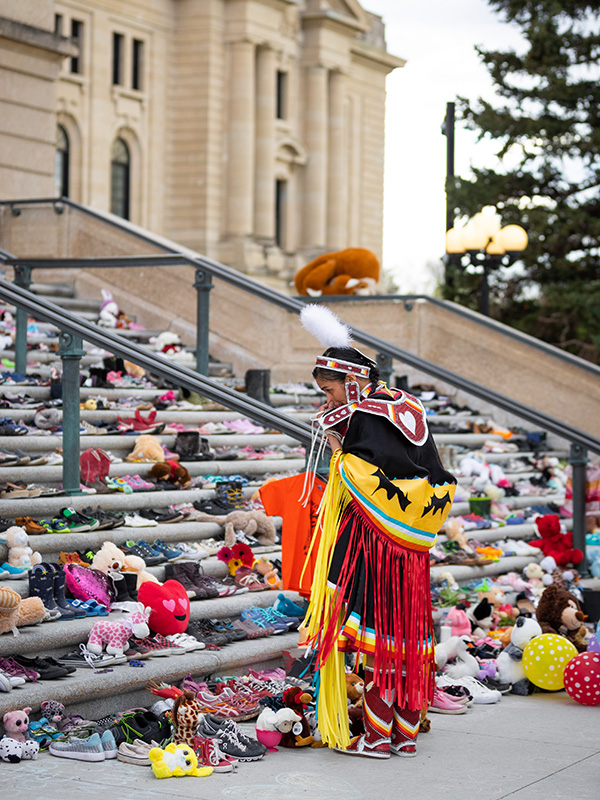 An Indigenous person dressed in indigenous regalia, looking at steps covered with children sized shoes