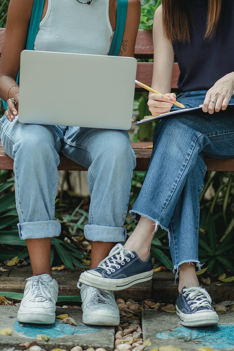 The legs of two women are seen as they sit on a part bench. Their legs are together and they have a laptop and notebook in their laps.