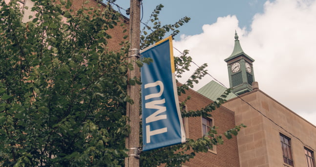 A blue TMU banner hanging on a lamp post in front of Kerr Hall's clock tower.