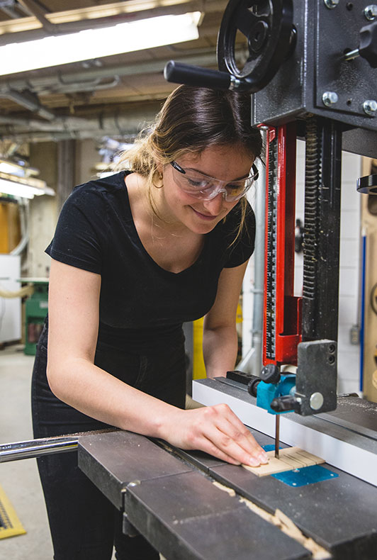 A student cutting a small piece of wood with a saw while wearing safety glasses.
