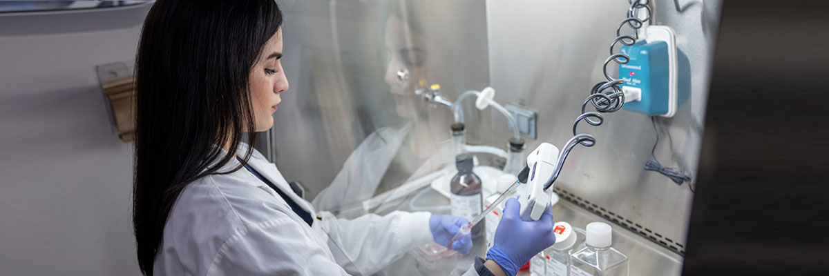 A female student in a white lab coat and gloves uses a fume hood.