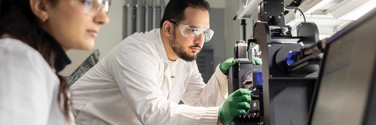 A male student makes an adjustment to lab equipment while a femals student reviews the equipment's computer screen. Both are wearing lab coats and safety glasses.