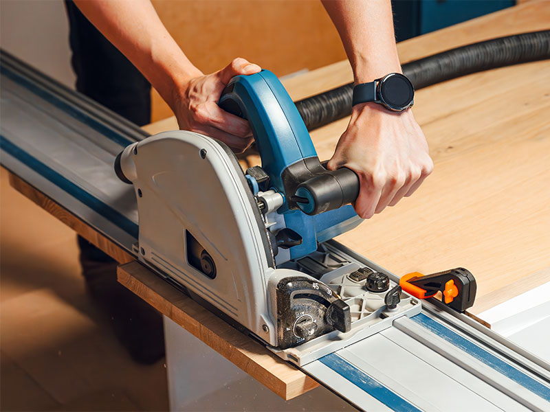 A pair of hands operate a circular saw to cut across a wide plank of wood.