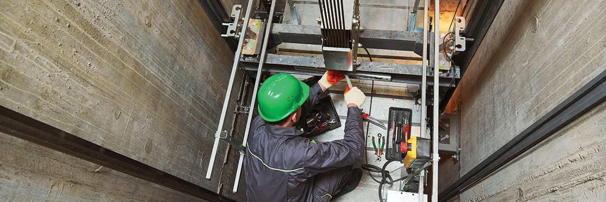 An elevator technician with a hard hat and insutrial PPE sits inside a concrete elevator shaft to work on cables. 