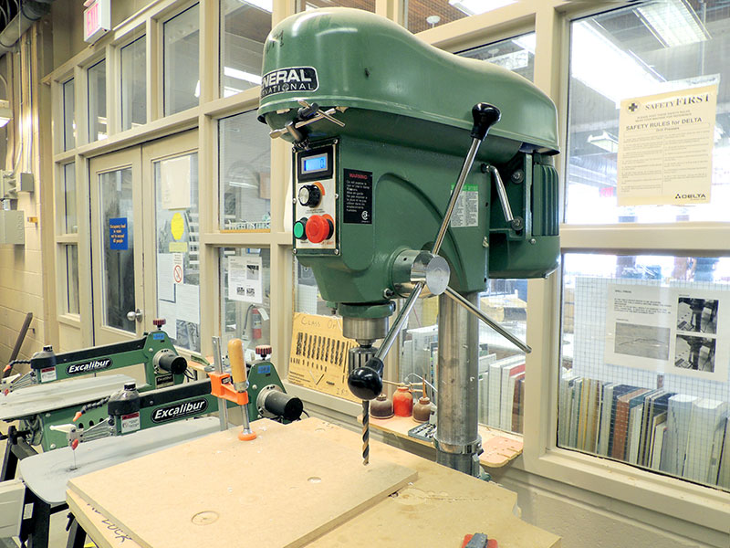 A drill press with a 3/8" bit ready to drill into plywood.
