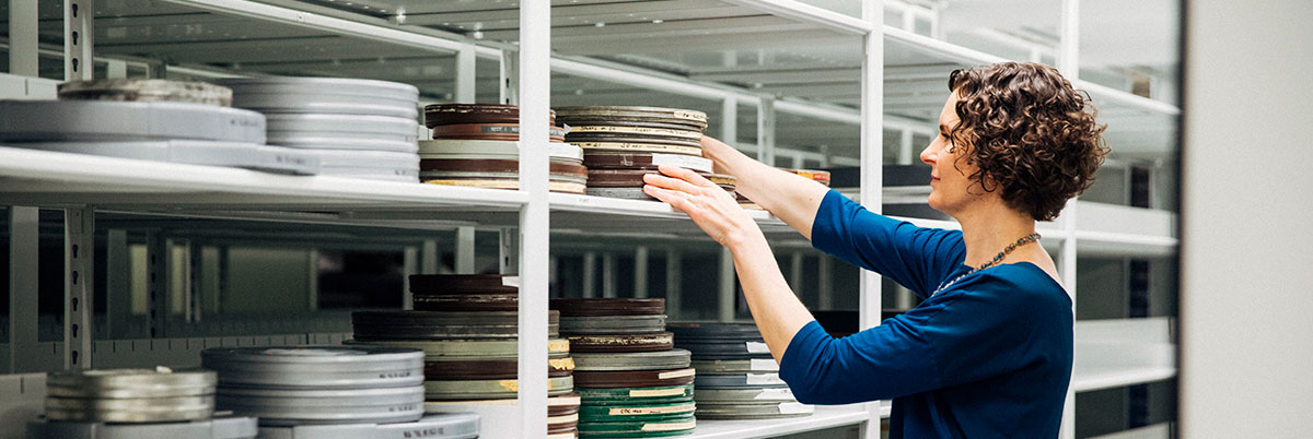 A female librarian reaches up high onto shelves to adjust film canisters in the library's film archive.