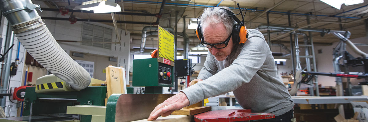 An employee uses a table saw to cut wood, while wearing safety gasses and sound-reducing ear muffs.