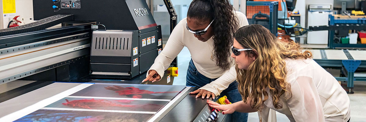 Two women wearing laser-safety glasses examine materials from a printer in the Creative Lab.