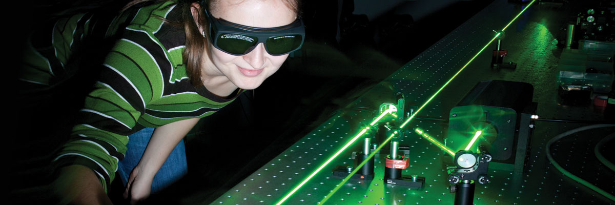 A female grad student wearing protective eyewear aims green lasers in a darkened lab.